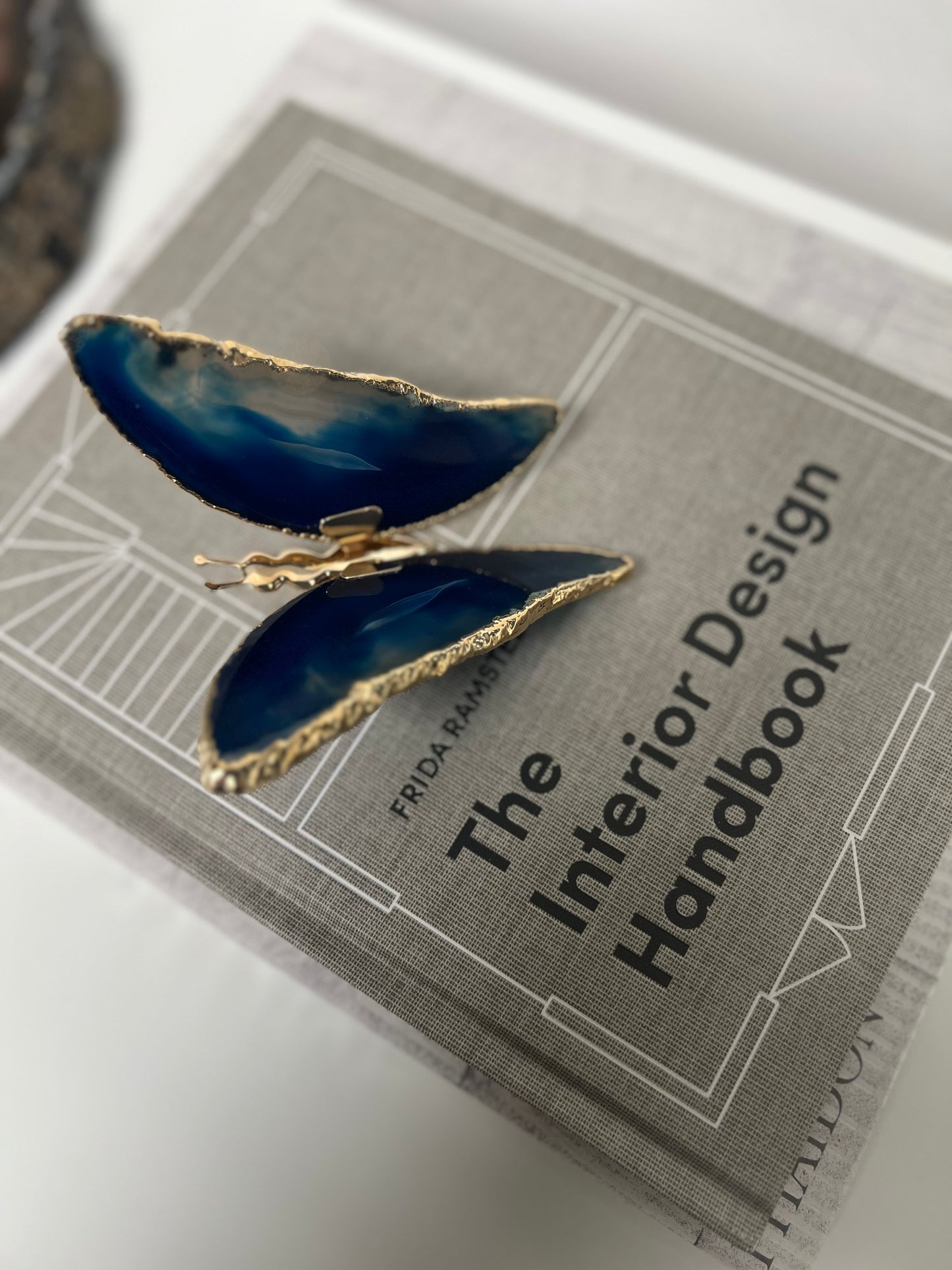Blue butterfly-shaped earring on a book titled 'The Interior Design Handbook'.