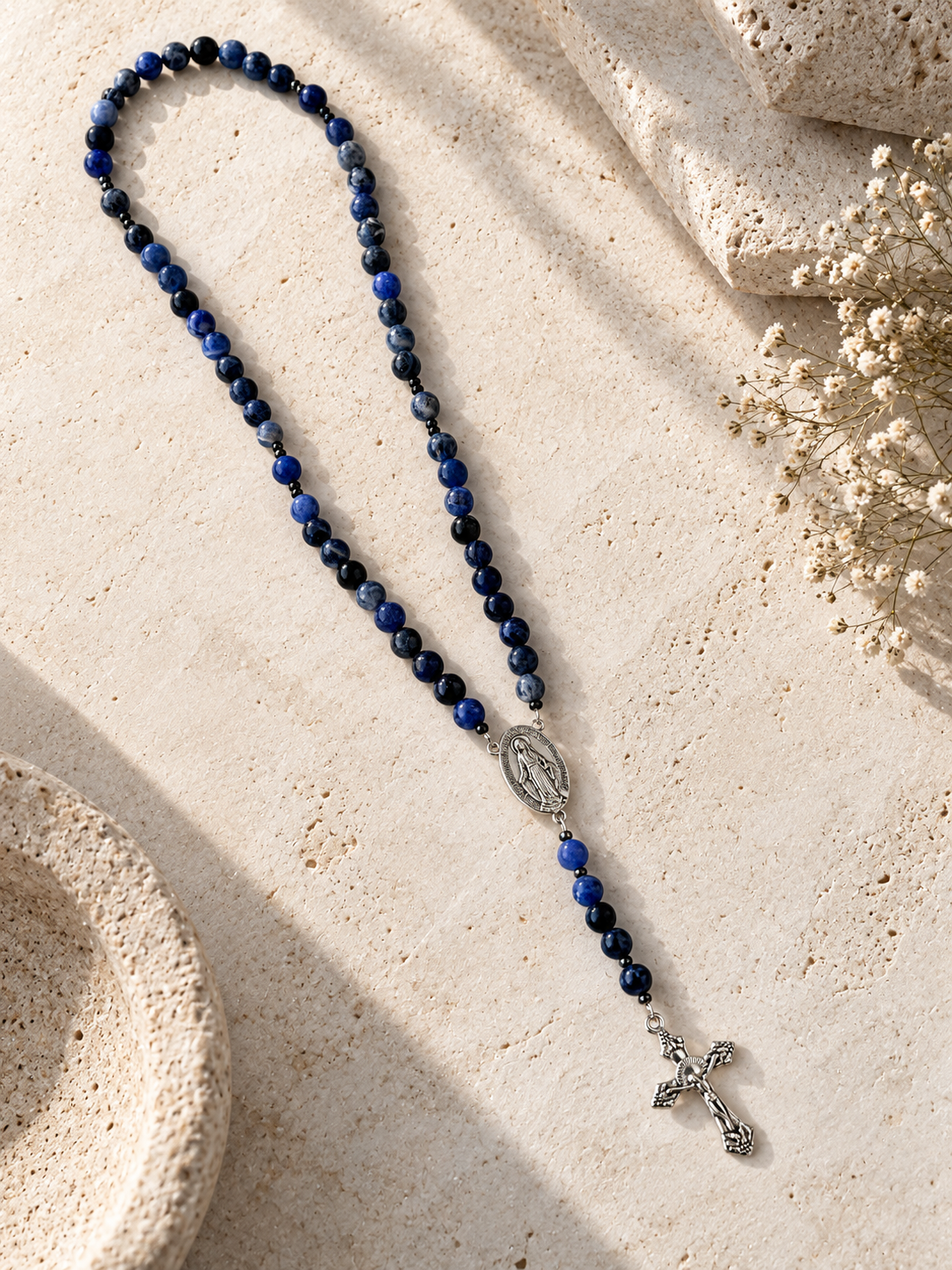 A beaded rosary necklace with dark blue and black round beads, a silver-toned Miraculous Medal centerpiece, and a dangling crucifix, arranged on a light beige stone surface with dried flowers and stone props.