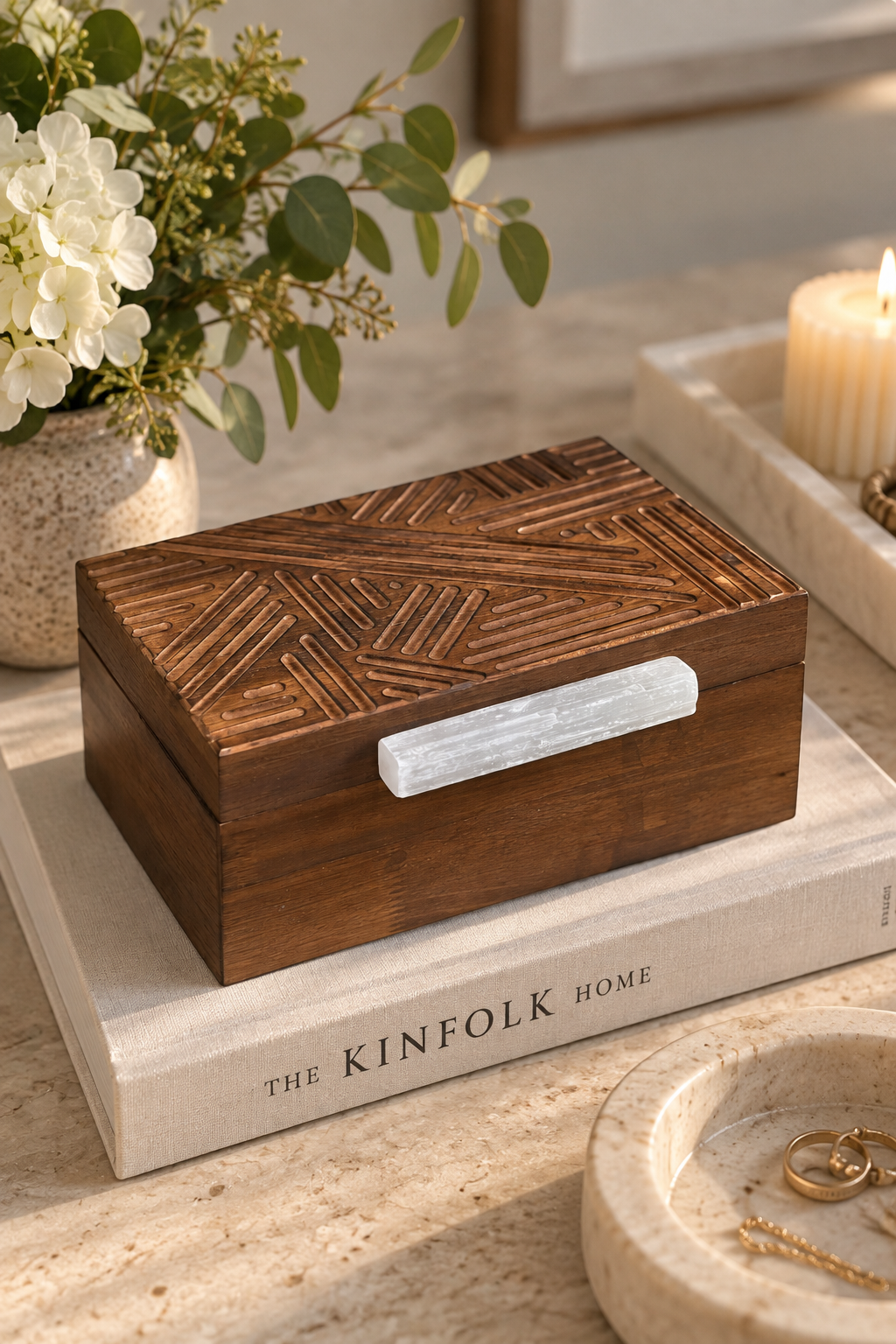A rectangular dark brown wooden keepsake box with an engraved geometric pattern on the lid and a white stone handle, displayed on a book in a styled home setting.