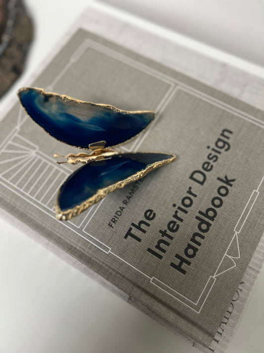 Blue butterfly-shaped earring on a book titled 'The Interior Design Handbook'.