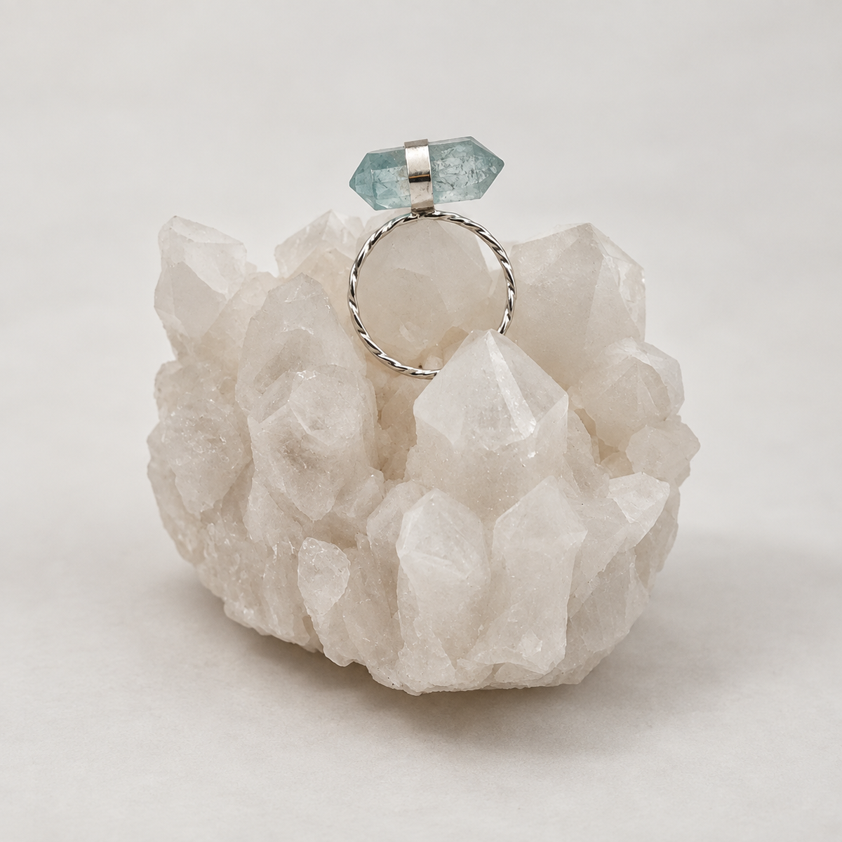 A silver-toned ring with a twisted band, a small silver cap, and a faceted light blue gemstone, displayed on a white crystal cluster against a light gray background.