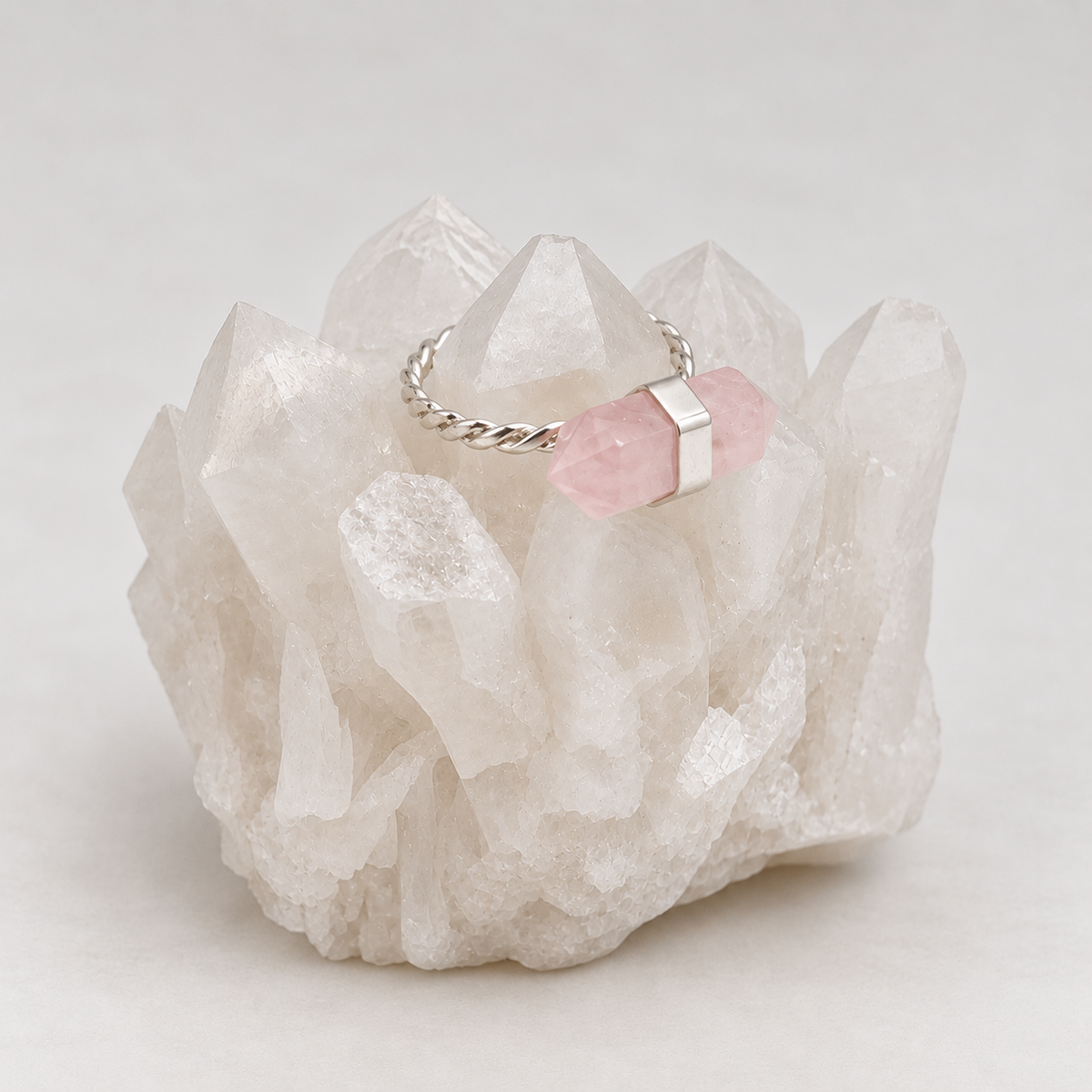 A silver ring with a twisted band and a pink stone set in a polished silver bezel, displayed on a white crystal cluster against a light gray background.