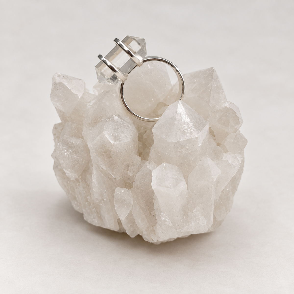 A silver ring with three clear faceted crystal-like gemstones, displayed on top of a white quartz crystal cluster against a light neutral background.