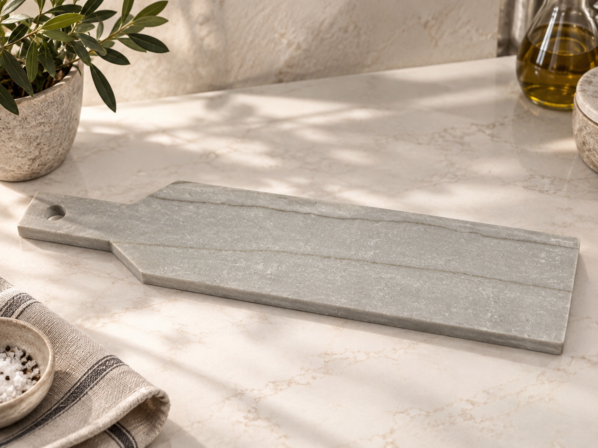 A rectangular gray stone serving board with a handle and a small round cutout, placed on a light marble countertop with a potted plant, bowl, and bottle in a styled kitchen setting.