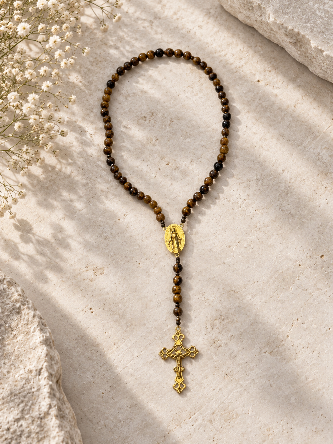 A beaded rosary necklace with brown and black tiger's eye beads, a gold oval medallion, and a gold cross pendant, arranged on a textured beige stone surface with dried white flowers.