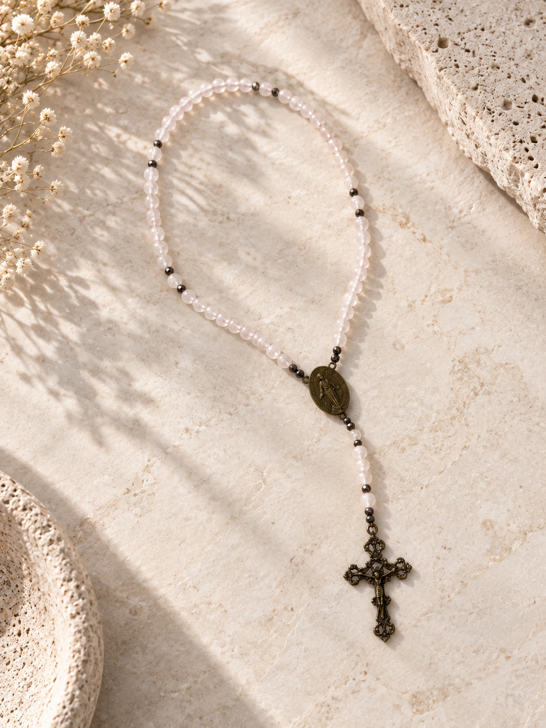A beaded rosary necklace with pale pink and white beads, small black spacer beads, a bronze oval medallion, and a dark bronze cross, arranged on a textured beige stone surface with dried flowers and soft shadows.
