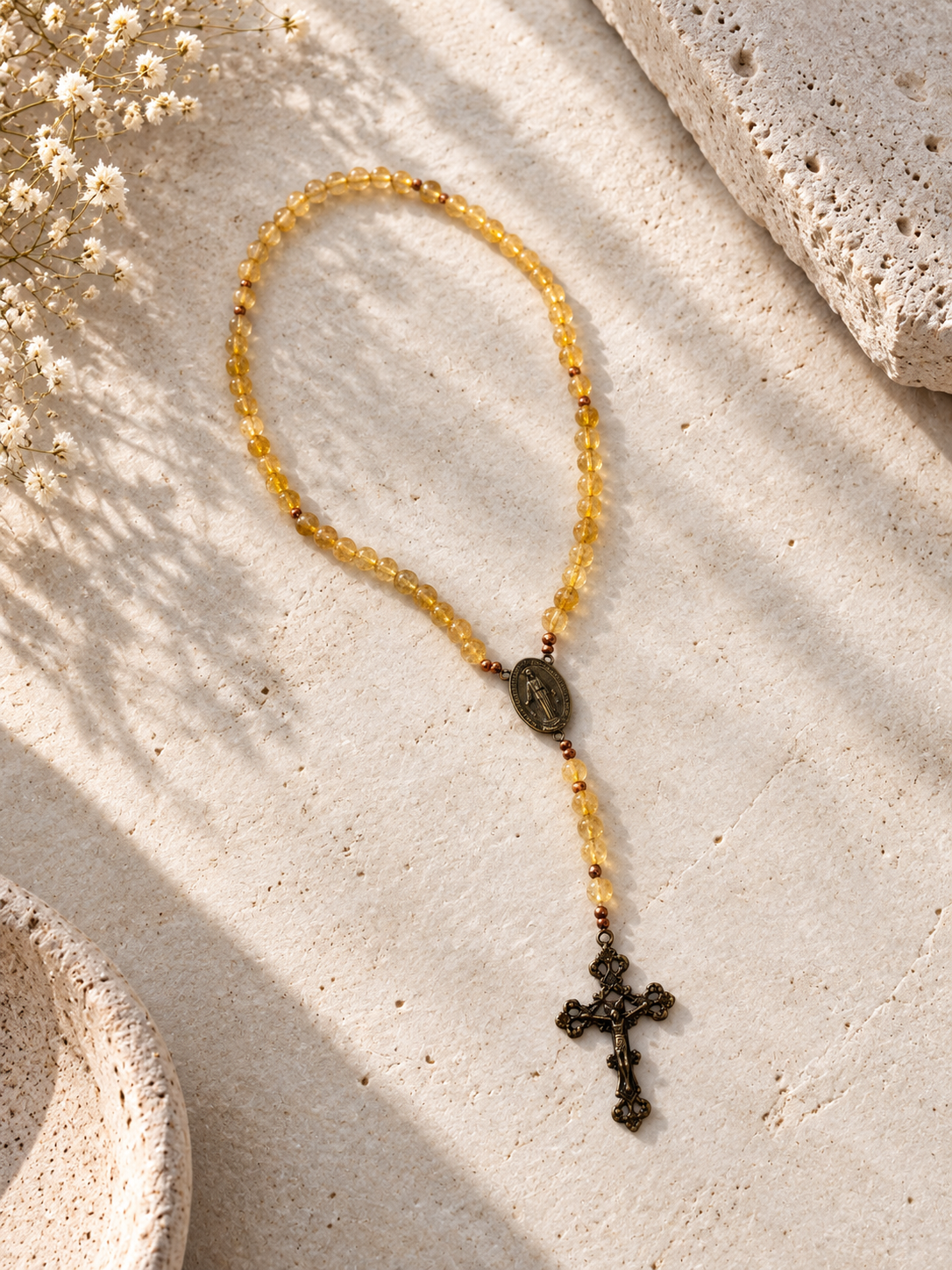 A beaded rosary necklace with translucent yellow beads, a small oval medallion, and an antique bronze cross, arranged on a textured beige stone surface with soft shadows.