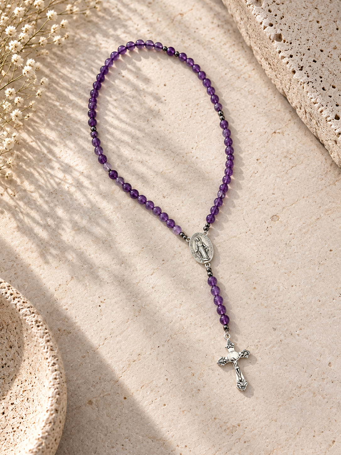A purple beaded rosary necklace with a silver Miraculous Medal and crucifix pendant, styled on a textured beige stone surface with dried flowers.