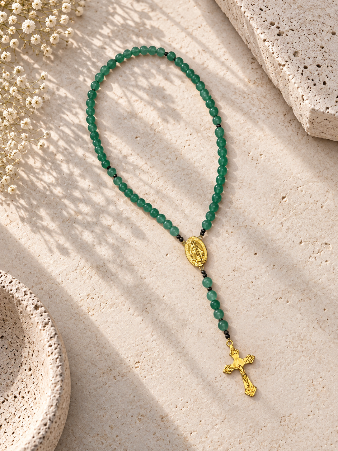 A green beaded rosary necklace with gold-toned religious medallions and cross, arranged on a light beige textured stone surface with soft shadows and small white flowers.
