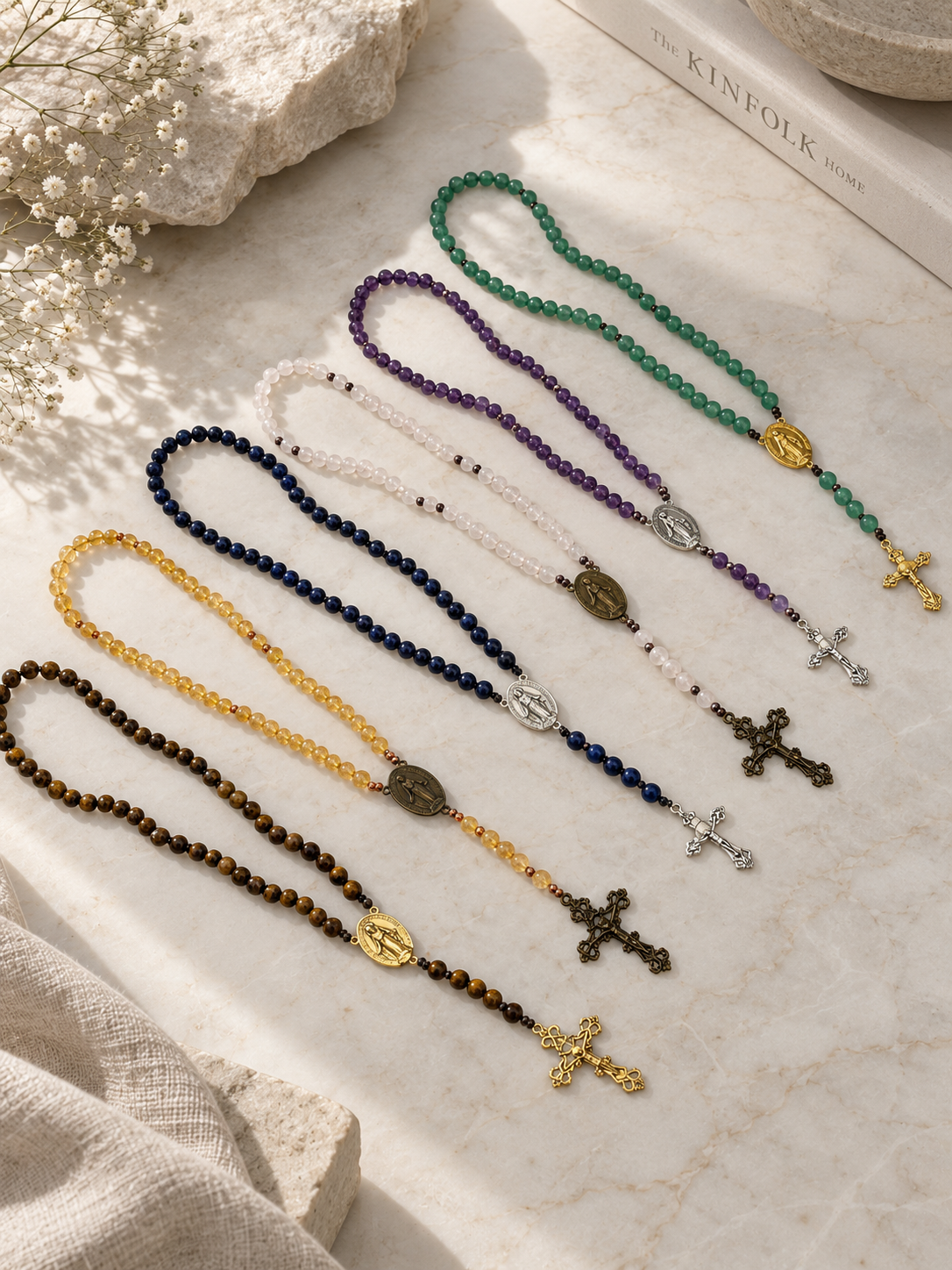 Six beaded rosary necklaces in different colors (green, purple, white, yellow, navy, and brown) are arranged diagonally on a light marble surface with linen and dried flowers as props.