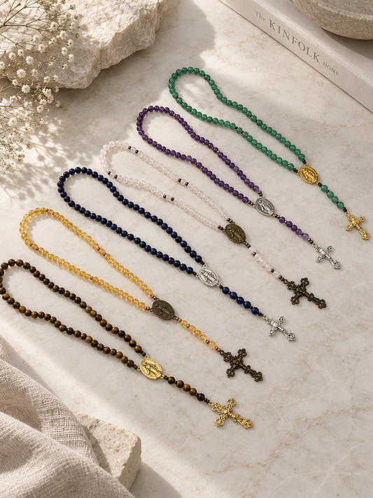 Six beaded rosary necklaces in different colors (green, purple, white, yellow, navy, and brown) are arranged diagonally on a light marble surface with linen and dried flowers as props.