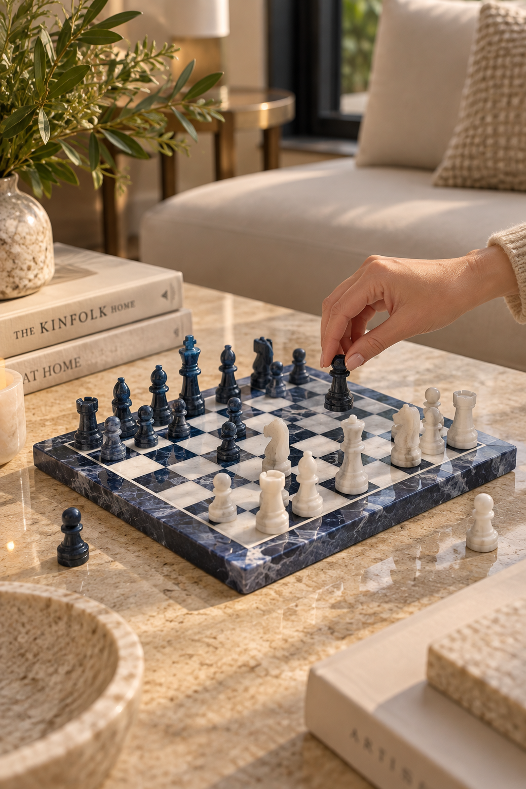 A marble chess set with dark blue and white pieces arranged on a stone board on a beige marble table in a modern living room. A person's hand is placing a black chess piece, with books, a plant, and a sofa in the background.